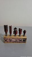 Six assorted router bits with red painted bodies mounted in a worn wooden holder, shown from front view.