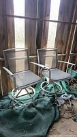 Two beige outdoor swivel chairs with metal frames and mesh fabric seats and backs. Chairs have armrests and swivel bases. Shown placed on green tarp inside a wooden enclosure.