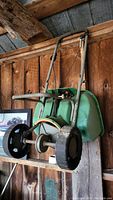 Side-angle view of a green lawn seed spreader showing large wheels, metal frame, and green hopper tank, hanging on wood wall.