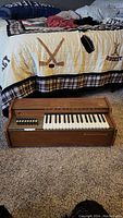 Image of vintage small piano keyboard on carpet with a bed in background. Shows full front of piano and part of keyboard.