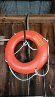 Full view of bright orange life ring float hanging against wooden background, with white rope attached.