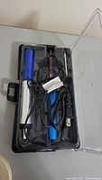 Top view of soldering iron kit in black plastic case with transparent lid, showing soldering iron handle colored blue and silver heating element, power cord, and metal accessories.