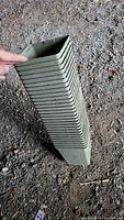 Stack of approximately 20 green square plastic plant pots held upright showing the square opening and sides in outdoor gravel ground environment.