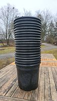 Side view of stack of approximately 20 black plastic plant pots stacked inside each other, showing ridged sides and slightly worn condition.