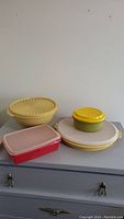 Photo showing five Tupperware containers: three round with lids (yellow and off-white), one small round with yellow lid, and one rectangular with pink lid.