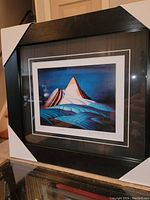 Framed print showing the mountain with blue foreground and white and orange peaks, with black frame and white corners protecting the frame.