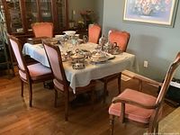 Six dining chairs around a wood dining table, covered with a white tablecloth and silver serving ware. The chairs have wooden frames and pink velvet upholstery, with two armchairs and four side chairs visible.