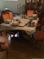 Full view of rectangular wood dining table covered partially with protective cloth and various silverware on top. The table is surrounded by upholstered chairs with reddish-orange cushions.