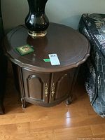Deilcraft dark wood octagonal drum table with brass handles, positioned between upholstered furniture and hardwood floor visible. Shows detailed edge scalloping and cabinet door design.
