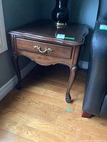 Side angle showing the wooden end table with one drawer, brass handle, curved legs, and carved details, positioned next to a dark couch on wooden flooring.