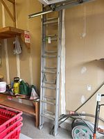 Photo of aluminum extension ladder standing folded near wall in garage with tag attached.