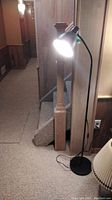 Full view of black steel floor lamp standing on carpet next to stairs and wooden walls, with light on.