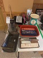 Overview photo showing various kitchen ware items on a white table including box grater, cookie cutters, loaf pans, flatware, marble cheese slicer, and metal tray with "Waffelbäckerei" design.