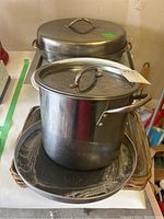 Large stainless steel stock pot with lid, oval roasting pan with lid, metal baking sheets, glass Pyrex and Corning Ware dishes with lids, and woven tray on garage table