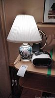 Desk lamp with floral ceramic base and white pleated lampshade next to white and black Sony Dream Machine clock radio on wooden surface