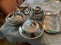 Wide view of all metal serveware items including covered bowls, tray, butter dish, gravy boat with spoon, and salt and pepper shakers arranged on table.