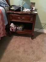 Nightstand positioned next to bed with drawer facing camera, showing carved wooden details, brass handle, and open shelf underneath. Items on top and under shelf not included.