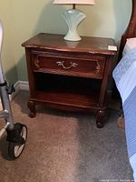 Front view of wooden nightstand with drawer pulled closed and lamp on top, showing decorative brass handle and carved leg detail.