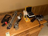 Photo showing the Kodak EK6 camera in original boxed packaging, Kodak Ektralite 10 flash, HP315 and Blacks cameras, and two tripods on a wood surface, cables not included.