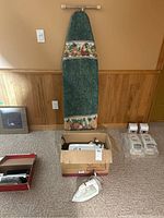 Ironing board standing against wall, white Braun iron on carpet floor, Kenmore sewing machine visible in box on floor along with other accessories.