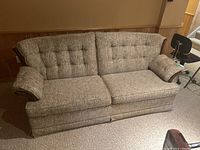 Front and side angled view of sofa bed showing tufted back and seat cushions, armrests with wood veneer trim, and tweed fabric upholstery in light brown and grey tones.