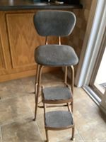 Photo showing full side view of folding metal step stool with padded seat and two padded steps covered in grey fabric.