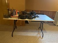 Folding table with metal legs and white laminate top, set up with various kitchen items on top in a basement room.