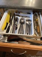 Drawer with Henkel stainless steel cutlery including forks, spoons, knives, wood-handled utensils, yellow-handled brush, and metal tongs.