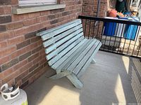 Front-left angled view of painted wooden bench against a brick wall on a concrete porch.