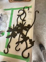 Three wrought iron decorative garden hooks laid on a white surface with a green tape marking '17'. Shows the full set with decorative details of bird, dragonfly, and flowers.