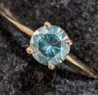 Close-up of 10K gold ring with round teal-blue moissanite gemstone set in four prongs on polished band.