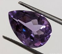 Close-up of genuine amethyst gemstone held by tweezers showing faceted teardrop cut and purple color