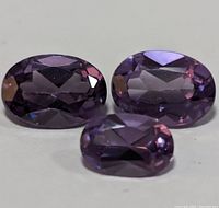 Three oval faceted genuine amethyst gemstones displayed on a white background in varying sizes showing clarity and purple color.