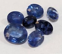 Seven loose natural blue sapphire gemstones arranged on a white background showing various faceted oval shapes and different blue color saturations.
