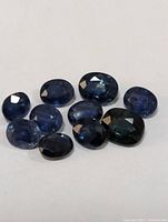 Photo showing nine loose blue faceted sapphire gemstones in various oval and round shapes on a white background.