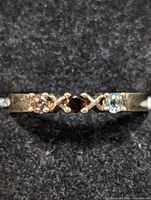 Close-up front view of silver band ring featuring three different colored gemstones: dark red center stone, light blue and light pink stones on sides with an 'X' design in between the stones.