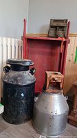Photo of large black milk can, silver milk can with wooden dispenser base, and red wooden box behind.