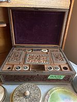 Carved Anglo-Indian sewing box with velvet lined lid, showing carved animal and floral motifs, several empty and occupied compartments below, and signs of wear and missing trim on lid and interior.