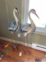 Both heron sculptures standing side by side on wooden floor beneath window