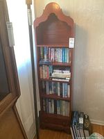 Front view of tall narrow wooden bookcase with various books on shelves and small drawer at bottom.