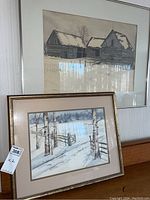 Photo showing both framed prints together on a shelf, one with barns in snow and one with snowy forest and fence.