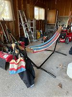 Wide photo of fabric hammock with striped colors on black metal stand inside garage. Includes storage bags hanging on stand.
