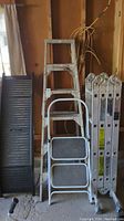 Photo of all four items: a black plastic folding ramp, a white two-step step ladder, an aluminum 6-foot A-frame ladder, and a foldable aluminum extension ladder, all standing against a wooden wall in a garage.