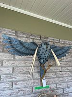Full view of blue-gray metal owl sculpture mounted on brick wall, wings spread, perched on a branch with attached light feather-like decorations.