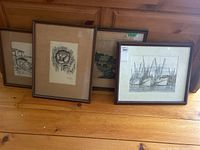 Multiple framed prints leaning against wooden wall showing various scenes including an owl, fishing boats, buildings, and landscapes.