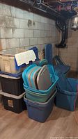 Stack of multiple plastic storage totes in black, blue, and beige color with some lids attached or open, located in basement corner with concrete block wall.