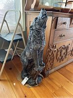 Front angled view of a standing wolf sculpture on a tree stump base, showing wolf looking upward howling, placed indoors by wooden furniture and a step stool.