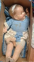 Full doll seated in box wearing blue and beige dress, showing signs of damage and stains.