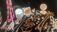 Full lot displaying two mirror/powder compacts, necklaces, bracelets, rings, charms, and brooches on black background