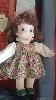 Vintage porcelain doll with white blouse, floral dress, green shoes and curly brown hair.